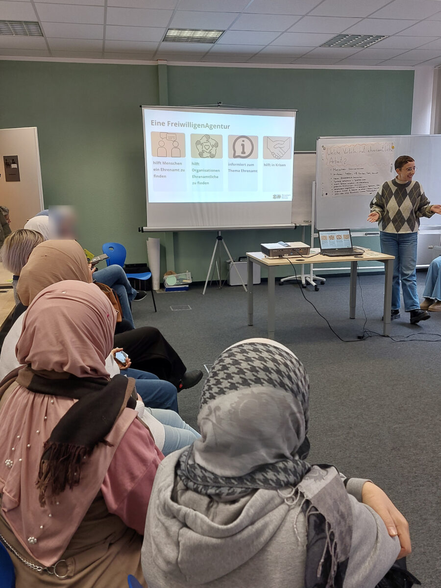 "Eine Frau hält einen Vortrag vor einer Gruppe von Menschen, die auf Stühlen in einem Seminarraum sitzen. Auf der Leinwand ist eine Präsentation mit der Überschrift 'Eine FreiwilligenAgentur' zu sehen. Die Folie beschreibt Aufgaben der Agentur, wie das Helfen beim Finden von Ehrenämtern und das Informieren über Ehrenamtsthemen. Die Teilnehmenden, teilweise mit Kopftuch, hören aufmerksam zu.