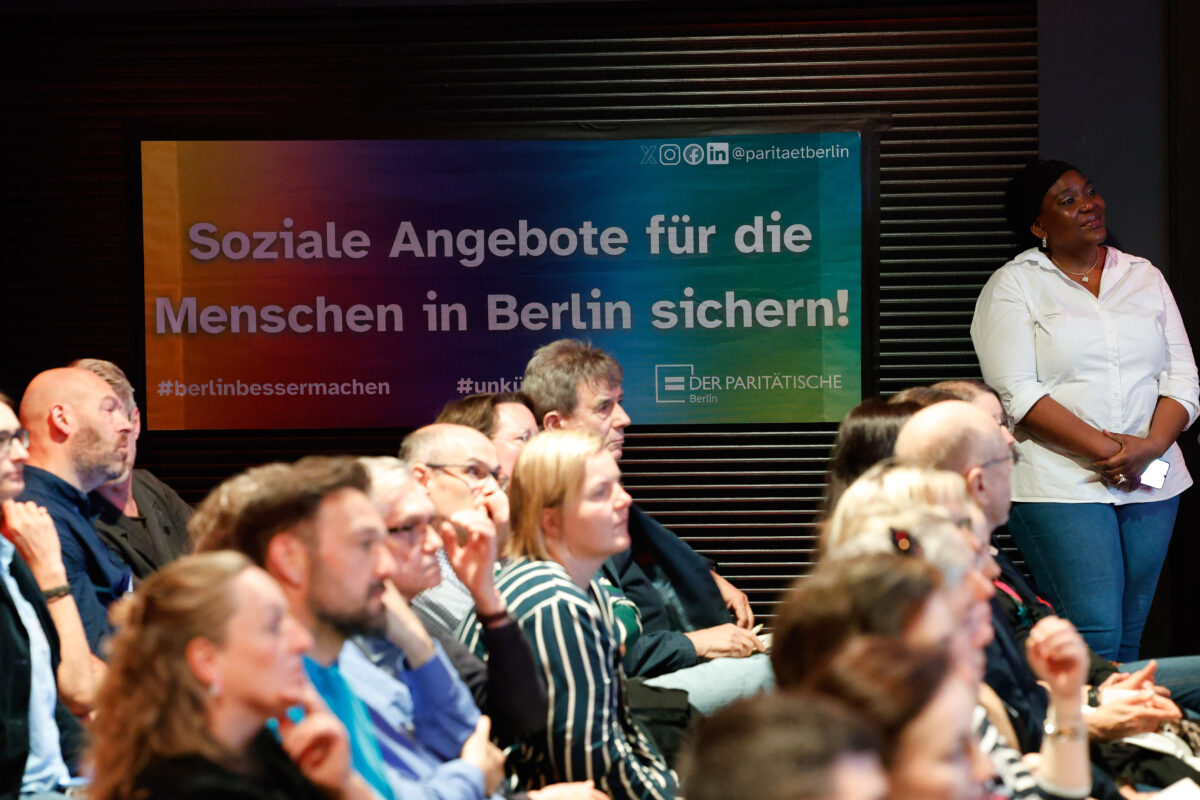 Publikum sitzt. Im Hintergrund ein Plakat mit der Aufschrift "Soziale Angebote für die Menschen in Berlin sichern!"